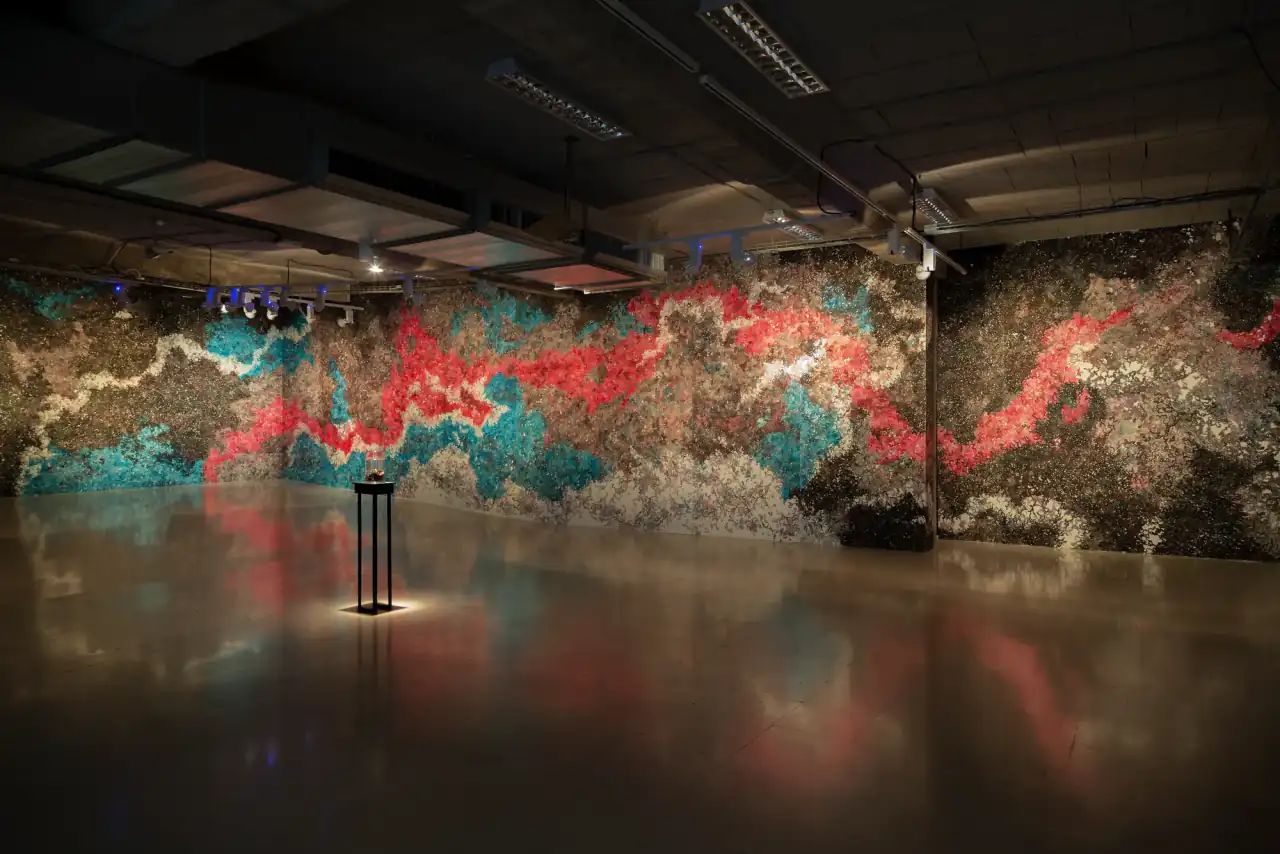 Sala de exposiciones en penumbra con dos paredes cubiertas de pintura sobre papel en rojo, turquesa y pardo, y una peana iluminada en el centro que se refleja en el suelo pulido.