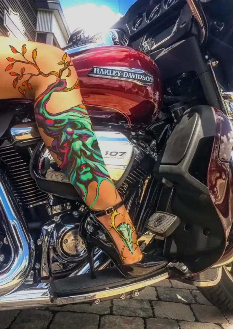 Diseño de María Vera mostrado en mockup sobre una fotografía de archivo: manga a color con una criatura verde, hojas naranjas y una gema azul, sobre una pierna apoyada en el motor de una Harley-Davidson roja.