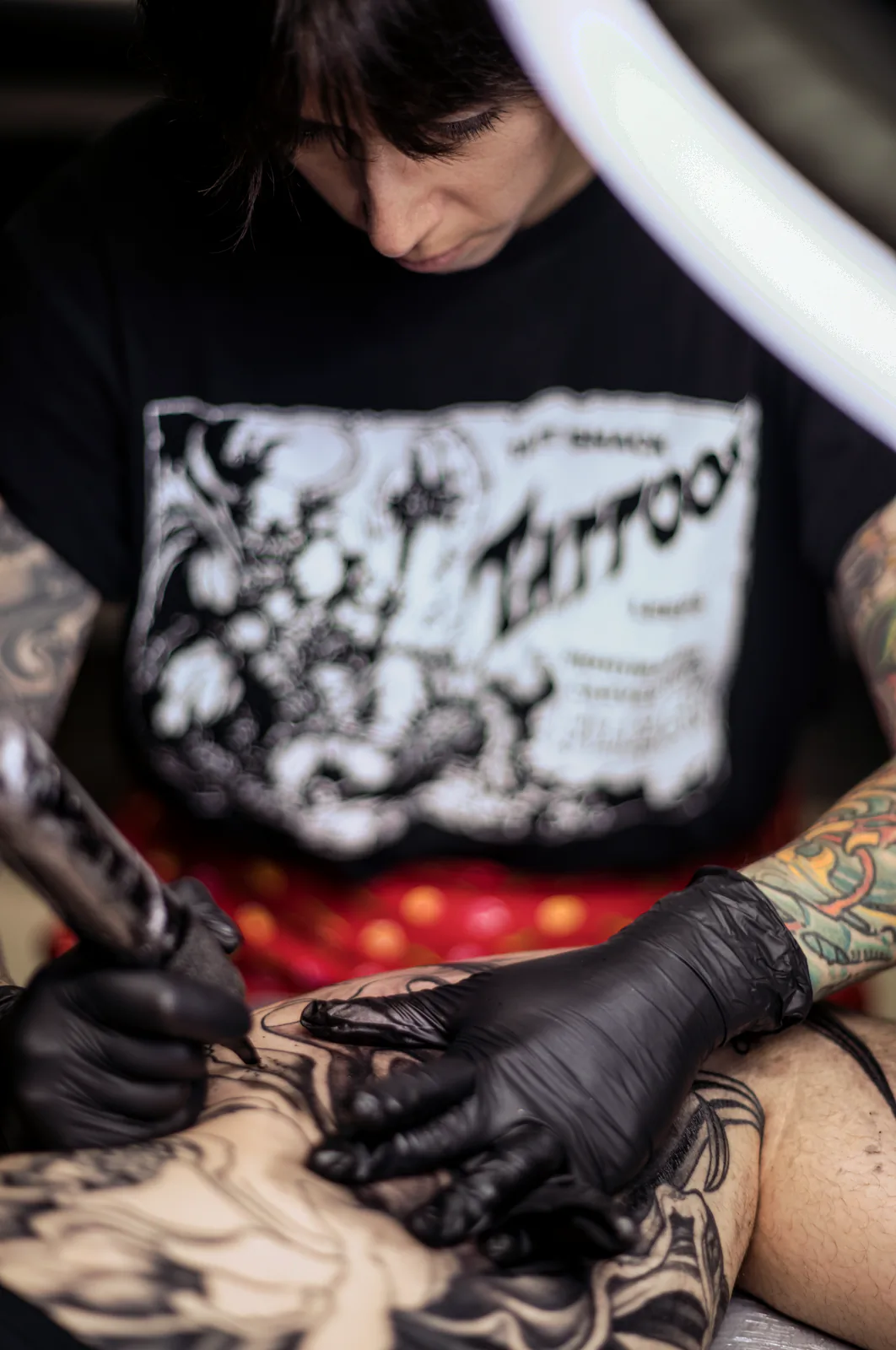 Vista cenital de una sesión: manos con guantes negros tatuando un muslo; camiseta negra con el rótulo «Tattoo».