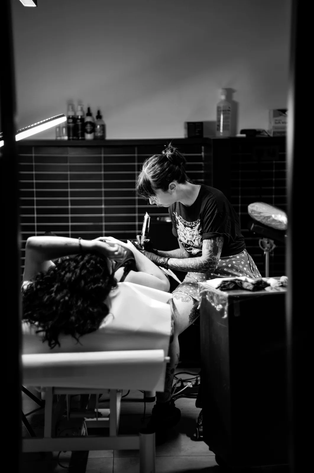 Tatuadora trabajando en la camilla junto al mueble del estudio, con luz lateral.