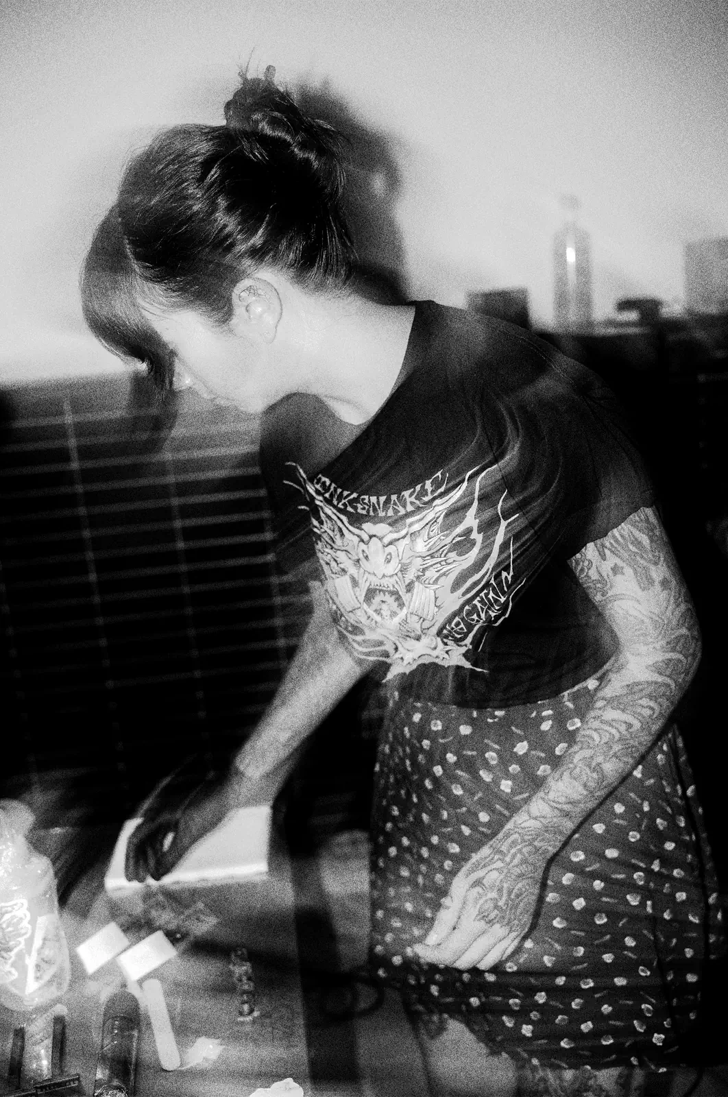 Retrato en blanco y negro de María, con moño y brazos tatuados, inclinada preparando el material sobre la mesa; camiseta de InkSnake, grano y barrido.