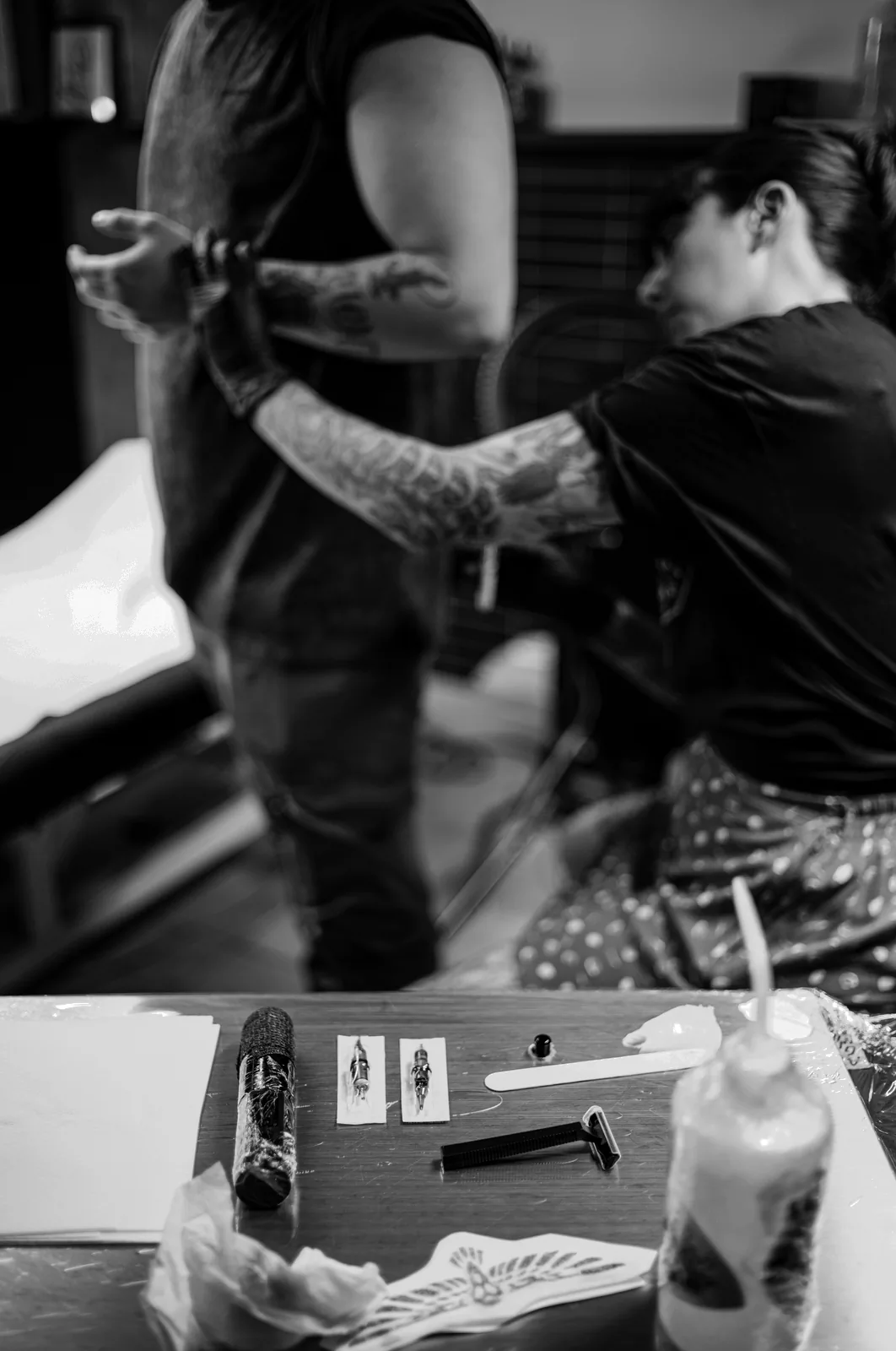 Tatuadora de pie tatuando el brazo de un cliente sentado; primer plano del utillaje en la parte baja.