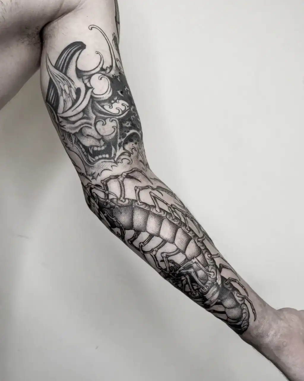 Manga en black & grey con una máscara hannya entre olas en el hombro y un ciempiés de placas bajando por el antebrazo, vista interior del brazo.